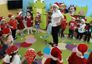 Grupa dzieci w czerwonych strojach i czapkach Mikołajów stoją w kole. Nauczycielka stoi na środku koła i pokazuje dzieci jak tańczyć.