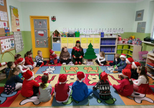 Grupa dzieci w czerwonych strojach i czapkach Mikołajów siedzą w kole i patrzą na panią przebraną za elfa.