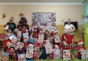 Grupa dzieci w czerwonych strojach i czapkach Mikołajów. Dzieci trzymają w dłoniach torebki prezentowe.