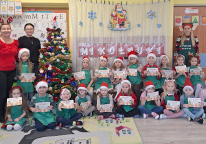 Grupa dzieci w czapkach Mikołajów i zielonych fartuszkach . Dzieci trzymają w dłoniach dyplomy.