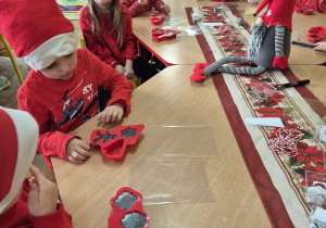 Grupa dzieci w czerwonych strojach i czapkach Mikołajów siedzą przy stolikach i wykonują mydełka.