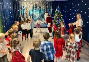 Dzieci w odświętnych strojach tańczą w kole z Świętym Mikołajem.