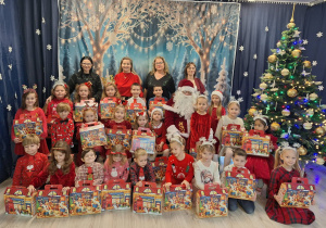 Dzieci w odświętnych strojach stoją i trzymają w dłoniach prezenty. Z tyłu stoją nauczycielki i Święty Mikołaj.