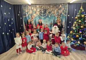 Dzieci w odświętnych strojach stoją i trzymają w dłoniach prezenty. Z tyłu stoją nauczycielki i Święty Mikołaj.