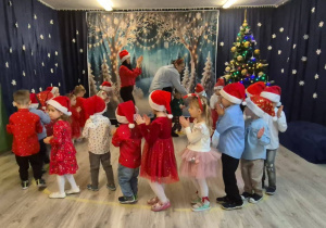 Dzieci w odświętnych strojach tańczą w kole z Świętym Mikołajem.