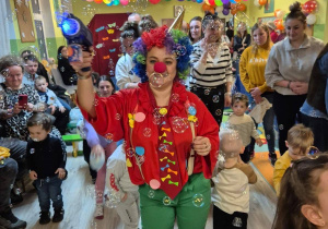 Pani przebrana w stój klauna, trzyma zabawkę, która wytwarza bańki mydlane. W tle małe dzieci oraz ich opiekunowie.