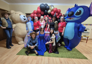 Grupowe zdjęcie pracowników przedszkola. Trzy kobiety siedzące z przodu, od lewej kowbojka, wróżka, piratka. Siedem kobiet od lewej kowbojka, pluszowy misio, stitch, meksykanka, księżniczka, myszka miki, czarownica, myszka minnie, dmuchany stitch. W tle ścianka dekoracyjna na stelarzu, czarne złote oraz czerwone balony.