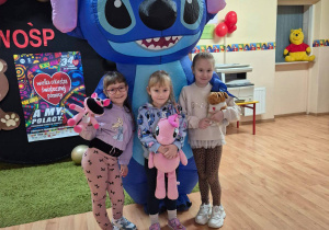 Zdjęcie przedstawiające trzy dziewczynki przytulające się do dmuchanego niebieskiego Sticha - postaci z bajki.