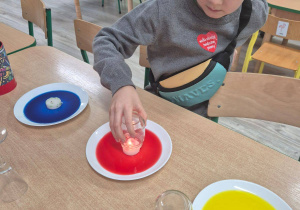 Zdjęcie przedstawiające chłopca trzymającego szklany pojemnik nad zapaloną świeczką, postawioną na talerzyku z wodą zabarwioną na czerwono.