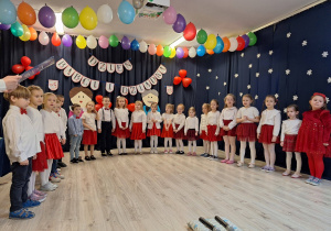Grupa dzieci stojąca podczas występu z okazji Dnia Babci i Dziadka. Dziewczynki ubrane w białe bluzki i czerwone spódnice, chłopcy w eleganckie koszule.