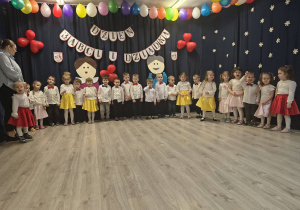 Grupa dzieci stojąca podczas występu z okazji Dnia Babci i Dziadka. Dziewczynki ubrane w białe bluzki, satynowe czerwone oraz żółte spódnice, chłopcy w eleganckie koszule.