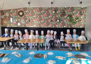 Grupa dzieci siedząca na ławce w czapkach kucharzy, przed nimi stoliki z ciastem do pizzy.