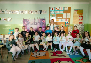 Grupa dzieci ubrana odświętne stroje. Dziewczynki trzymają w dłoniach kwiaty. Z nimi na zdjęciu Pani.