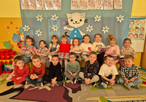 Grupa dzieci, dziewczynki siedzące na ławce trzymające w ręku upominki, na dole chłopcy. W tle dekoracja Dzień Kobiet wizerunek postaci "Kicia Kocia".