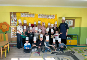 Grupa dzieci w czapkach kucharzy stojąca na tle dekoracji, wraz z nimi dwie Panie oraz Pan.