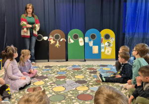 Pani prowadząca warsztaty, cztery kosze oraz przyklejone śmieci posegregowane. Dzieci siedzące na macie słuchające wykładu.