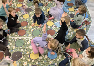 Dzieci bawiące się na macie z kółeczkami w kolorach, kładą ręce i nogi na odpowiednich kolorach.