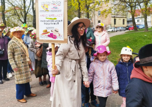 Kobieta stojąca w kapeluszu, trzyma w dłoniach tabliczkę z plakatem. Obok stoi dziewczynka w kapeluszu