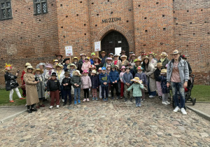 Grupa dzieci i i seniorów w kapeluszach stojąca na tle zamku łęczyckiego.