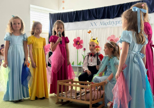 Grupa dzieci stojąca nad kołyską z lalką. Dziewczynki przebrane w dworskie stroje. Dwoje dzieci przebranych za króla i królową siedząca przy kołysce.