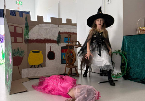 Czarownica siedząca nad wrzecionem. Dziewczynka leżąca w stroju królewny. W tle zamek z kartonu.