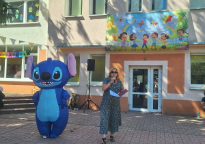 Maskotka Stitch, obok kobiety. Pani dyrektor przedszkola trzyma mikrofon do którego mówi na tle przedszkola.