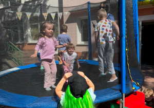 Czworo dzieci skaczących na trampolinie, dziewczynka w zielonych włosach stojąca tyłem patrzy na nie.