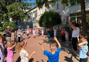 Grupa dzieci stojąca w kole z dwoma opiekunkami. Trzymają ręce w górze, tańczą. W tle dzieci skaczące na trampolinie.