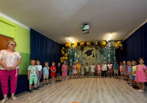 Grupa dzieci stojąca ze swoją Panią z lewej strony, śpiewająca piosenki. Wszyscy ubrani kolorowo na tle dekoracji absolwentów