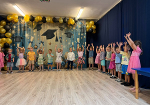 Grupa dzieci stojąca i śpiewająca piosenki. Wszyscy ubrani kolorowo na tle dekoracji absolwentów