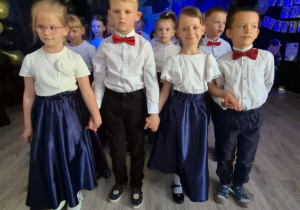 Dzieci tańczące poloneza, trzymają się za ręce, dziewczynki unoszą jedną ręką swoją spódnicę. Na pierwszym planie dwie pary obok siebie.