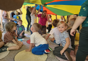 Dzieci siedzą na dywanie i uśmiechają się nad nimi kolorowa chusta animacyjna