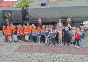 dzieci i nauczycielki w kamizelkach odblaskowych stoją na tle wojskowego samochodu ciężarowego