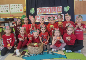 Dzieci ubrane na czerwono z opaskami w kształcie jabłka siedzą na tle dekoracji w jabłka,