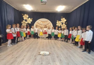 Grupa dzieci odświętnie ubrana. Chłopcy na galowo a dziewczynki w białych bluzkach i kolorowych spódniczkach.