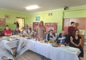 Odświętnie ubrani mężczyźni i kobiety siedzące przy zastawionym słodkościami i owocami stole.