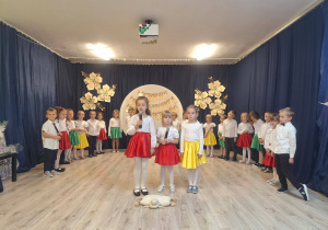 Grupa dzieci odświętnie ubrana. Chłopcy na galowo a dziewczynki w białych bluzkach i kolorowych spódniczkach. Na pierwszym planie trzy dziewczynki mówią do mikrofonu.
