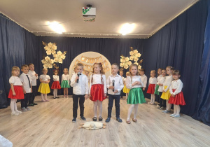 Grupa dzieci odświętnie ubrana. Chłopcy na galowo a dziewczynki w białych bluzkach i kolorowych spódniczkach. Na pierwszym planie dwie dziewczynki i dwóch chłopców mówią do mikrofonu .