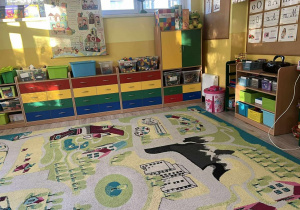 Na pierwszym planie dywan w jednię. Na drugim palnie są kolorowe szafki. na szafkach są pudełka z zabawkami.