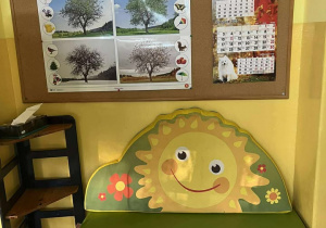 na zdjęciu widoczna jest półka z zabawkami na niej stoi materac z rysunkiem słonka. Nad szafka jest tablica z obrazkami pór roku.