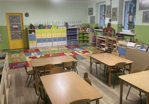 Sala przedszkolna grupy Misie, na pierwszym planie stoliki z krzesełkami, na drugim biurko nauczyciela, półki z zabawkami oraz książkami