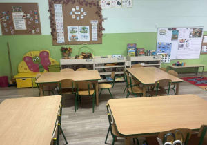 na pierwszym planie stoliki z krzesłami, na drugim półki z zabawkami oraz kolorowe tablice