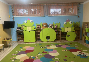 Sala grupy Żabki, kolorwy dywan z wizerunkiem gąsiennicy, w tle zielone szafki, zabawki, oraz telewizor na ścianie